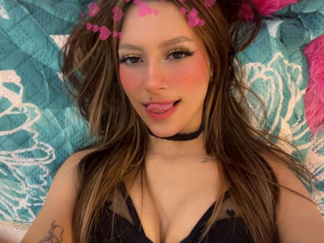 AfroditaWild has long brown hair, a pink flower headband, black choker, and is licking her pink-tinted lips. She's wearing a black lace top, showing her tits.