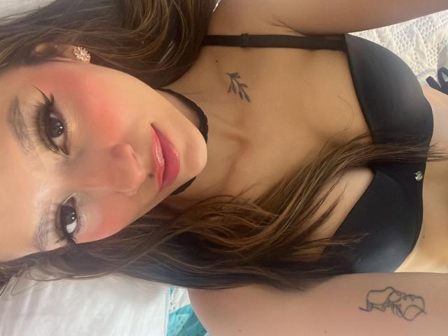 AfroditaWild with pink blushed cheeks, black bra, glossy pink lips, long brown hair, black choker, tattoos on chest and arm, looking at camera. Boobs in black bra.
