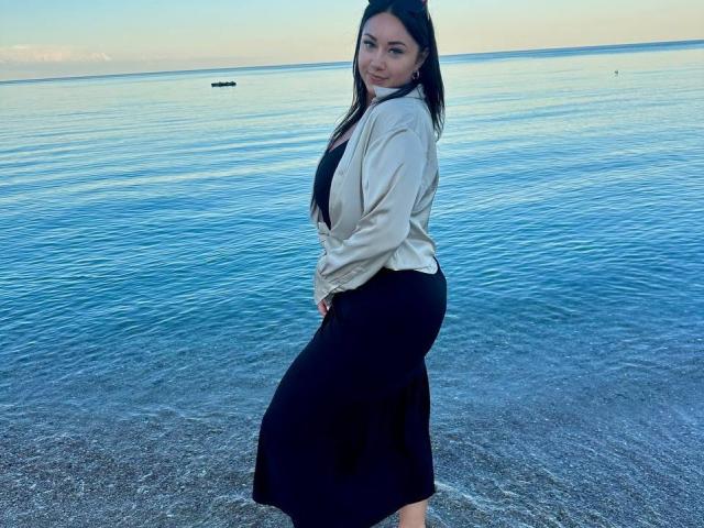 AdriannaGoldis stands by the water, wearing a beige shirt and black skirt, looking over her shoulder with a smile, showing her ass slightly. Her tits are small and cute.