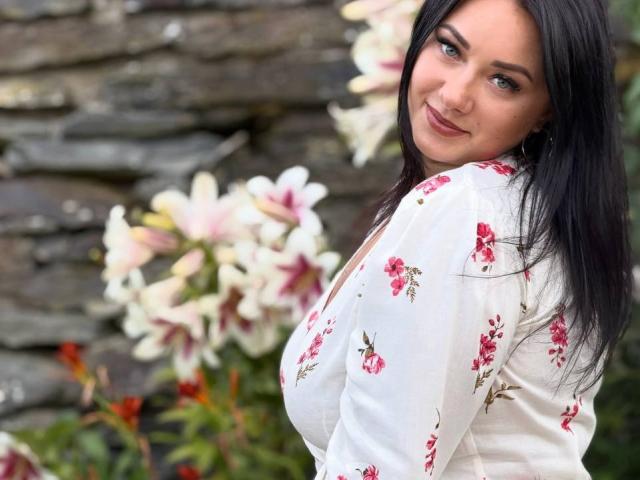 AdriannaGoldis with long black hair, smiling at the camera, wearing a white floral top, showing her boobs, standing near white and pink lilies.