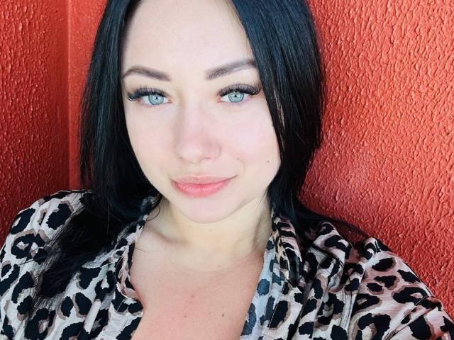 AdriannaGoldis has pale skin, blue eyes, and black hair. She's wearing a leopard print shirt, with full lips and a subtle smile. Her tits are not visible.