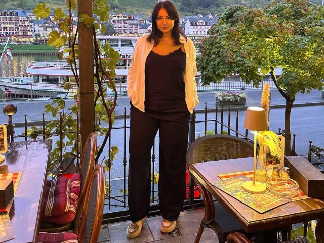 AdriannaGoldis stands with hands in black pants, white jacket, black top, and white sneakers, smiling at a river view from a patio with wooden table and lamp.