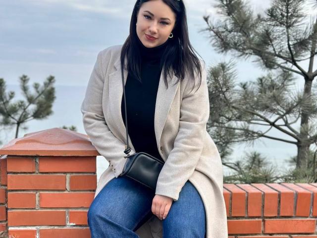 AdriannaGoldis with black turtleneck, beige coat, blue jeans, and black crossbody bag, sitting on red brick wall, holding her coat, with trees in background, smiling slightly.