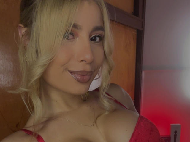 AmaraRicci, loira, com cabelo ondulado, usando um sutiã vermelho e um colar dourado, com a língua de fora, sorrindo, mostrando seios médios, pele clara e maquiagem discreta, em um quarto com pouca luz..