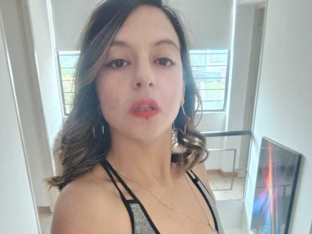 ArianaBlonde with wavy brown hair, red lipstick, and hoop earrings, wearing a gray and black halter top, looking at the camera. Her tits are small and she's cute.