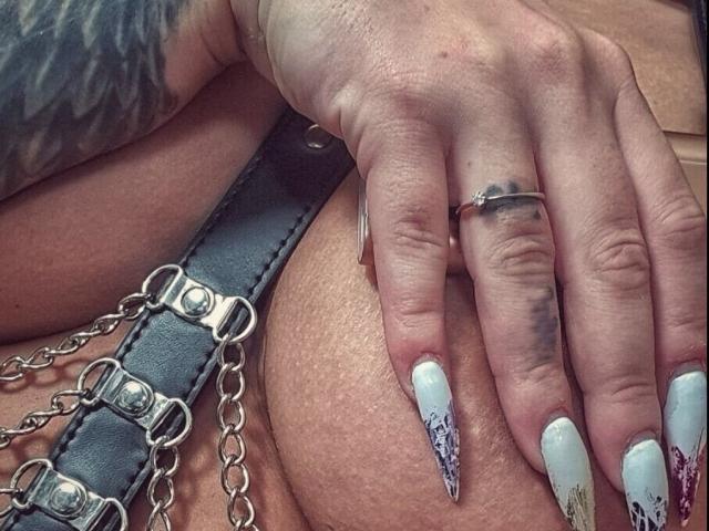 JustIzabel's tattooed hand with long, white, blood-splatter nails rests on her big ass. She's wearing a black leather belt with metal chains and a silver ring.