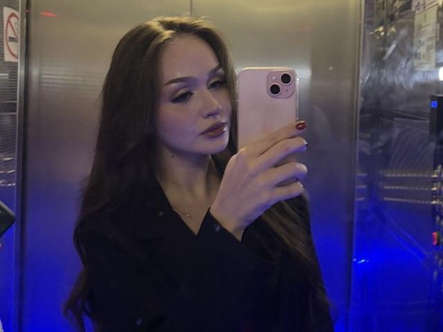 TamraBroxterman with long brown hair, wearing a black shirt, taking a selfie in a mirror, holding a pink iPhone, blue lighting, looking at the phone with a slight smile.