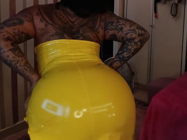 Chili with tattooed arms and back, wearing a shiny yellow latex dress, has a big, round ass, standing with hands on hips, in a dimly lit room.