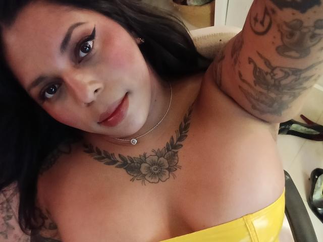Chili with black hair, tattoos, and pink eyeliner, wearing a yellow top, showing her large tits and flower chest tattoo, smiling softly, taking a selfie.