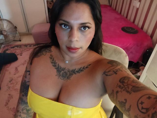 Une femme avec de grandes seins, vêtue d'un haut jaune sans bretelles, avec des tatouages sur la poitrine et les bras, des cheveux foncés, du rouge à lèvres, assise sur une chaise et regardant la caméra..