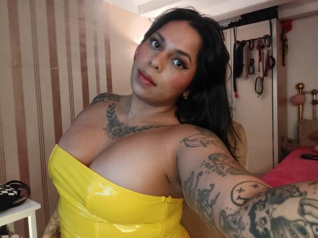 Chili with black hair, yellow strapless top, tattoos on arms and chest, and large boobs, taking a selfie in a room with striped wallpaper. Her eyes are blue.