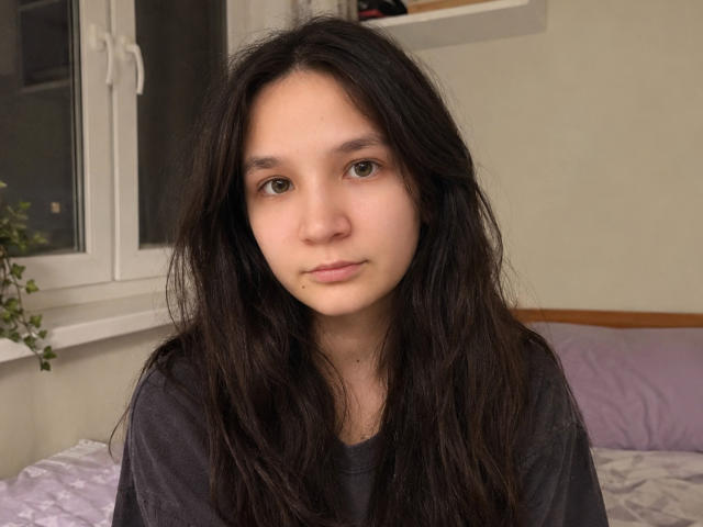 OdeliaHolen with long, messy black hair, wearing a dark shirt, has a neutral expression, brown eyes, and a small piece of food on her lower lip.