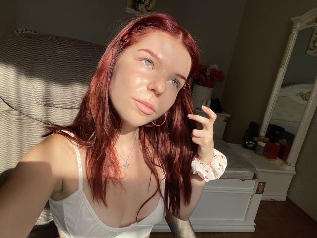 AlexXas, avec ses cheveux roux, sa peau claire et ses yeux verts, portant un débardeur blanc, des boucles d'oreilles cerceaux et un élastique à cheveux, assise au soleil, montrant son visage et sa partie supérieure du torse..
