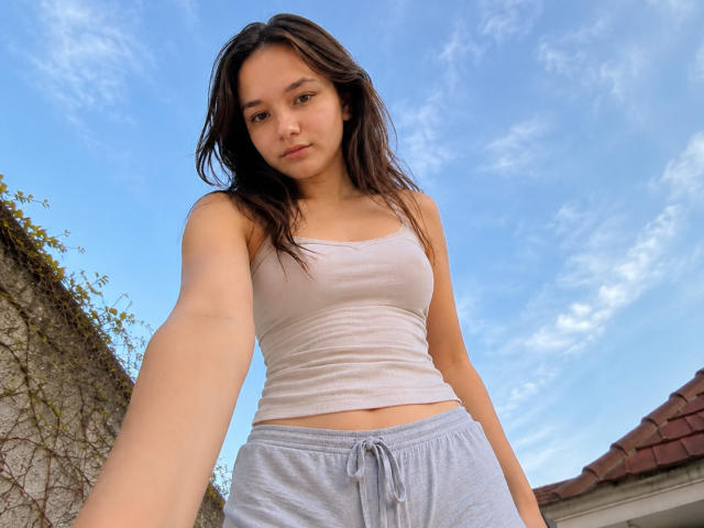 OdeliaHolen with medium tits in a white tank top and grey sweatpants, taking a selfie from a low angle, against a blue sky with wispy clouds.