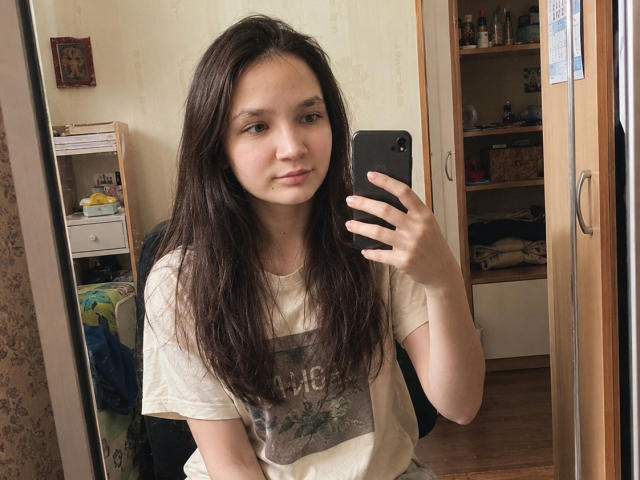 OdeliaHolen with long dark brown hair, wearing a beige t-shirt, holding a black phone, taking a mirror selfie, showing a slight smile, and a calm expression.