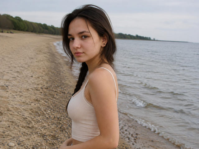 OdeliaHolen with dark hair in a braid, wearing a white tank top, showing her small tits, standing on a pebbly beach with a calm, grayish water background.