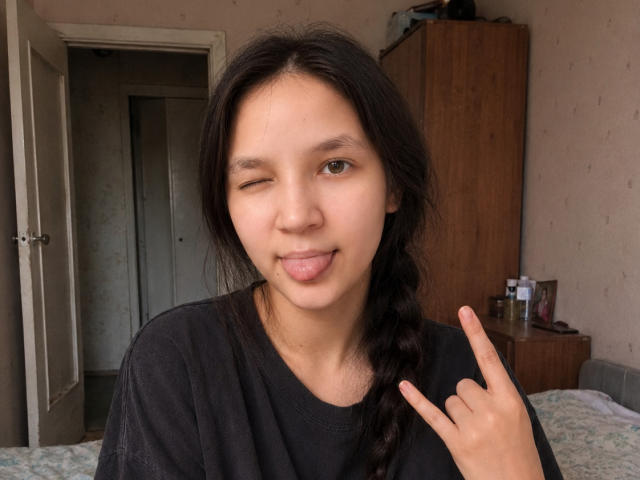 OdeliaHolen with long black hair in a braid, winking, sticking out her tongue, and giving a peace sign with her right hand, wearing a black shirt.