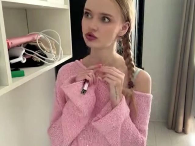 LawanaDavito with blonde braided hair, pink off-shoulder sweater, holding a pink hairdryer, tattoo on right hand, looking to the side, standing in front of a white shelf.