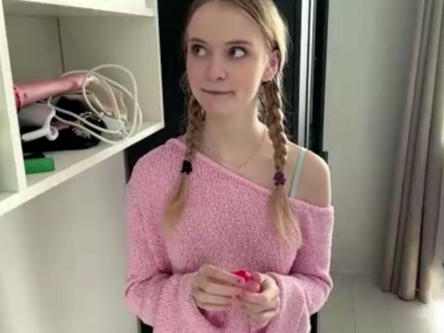 LawanaDavito with braided hair, pink off-shoulder sweater, holding pink nail polish, looking to the side, in a room with a white shelf and pink hairdryer.