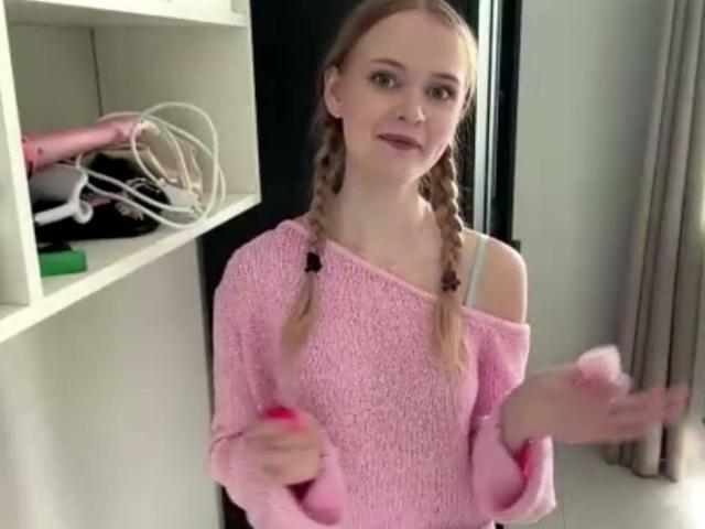 LawanaDavito with blonde braids wearing a pink off-shoulder sweater, smiling, hands raised, pink nail polish, white bra strap visible, pink hairdryer on white shelf in background.