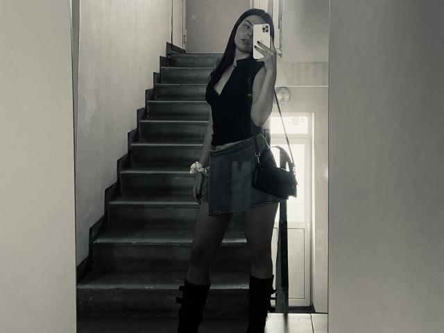 AlexXas, avec de longs cheveux foncés, un haut noir sans manches, une jupe en jean, des bottes montantes, tenant un téléphone, debout sur les escaliers, selfie devant un miroir, sac à main noir, élastique blanc au poignet..