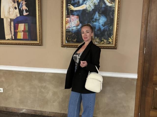 MysteryLaddy has tits, wearing a black blazer, blue pants, and a white bag, standing in front of paintings, looking at the camera, with blonde hair in a ponytail.