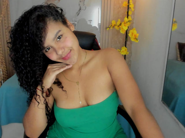 AriaVega with curly black hair, green strapless dress, small tits, gold necklace, smiling, hand on cheek, yellow orchids, sitting in chair, blue cloth, soft light.