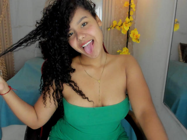 AriaVega with curly black hair, green strapless dress, gold necklace, tongue out, holding hair, smiling, brown skin, medium tits, yellow orchids in background.