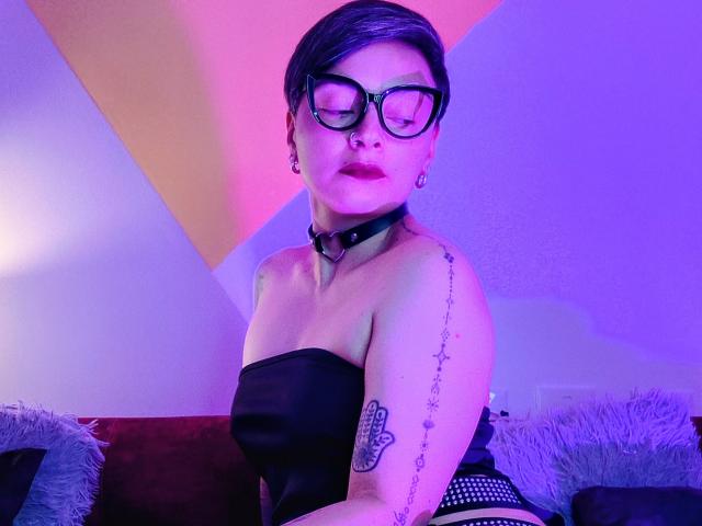 KatParadise, con cabello negro corto, gafas y un piercing en la nariz, vestida con un top negro sin tirantes y un collar de cuero, sentada en un sofá, mostrando un tatuaje en su brazo..