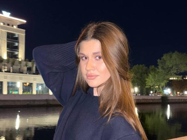 LakeishaCresencio with long brown hair, wearing a dark blue sweater, raising her right arm, has a serious expression, standing by a reflective water surface at night.