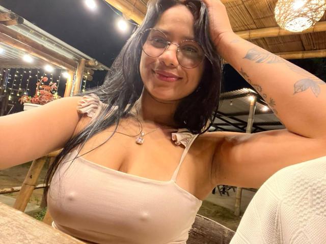 AlanaLuxe with glasses, smiling, wearing a tight beige top showing her tits, hand in hair, tattoo on arm, night setting, wooden roof, soft lights, warm skin.