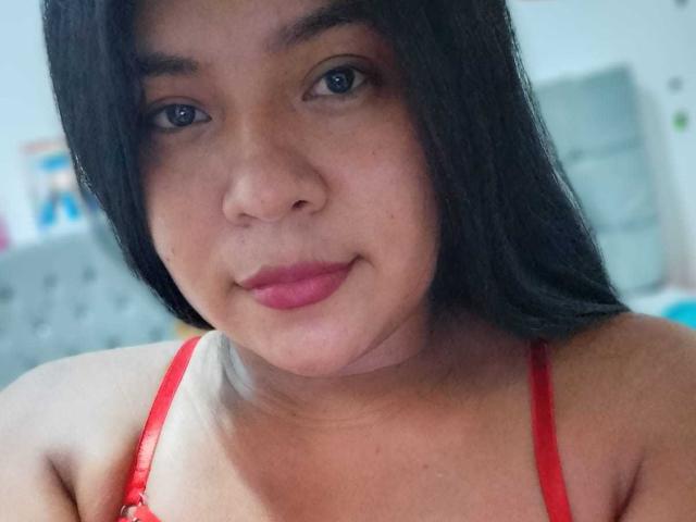 DulceChanell tiene una piel marrón media, cabello negro largo y lápiz labial rojo. Lleva un sostén rojo, y sus pequeños senos son ligeramente visibles. Está sonriendo suavemente..