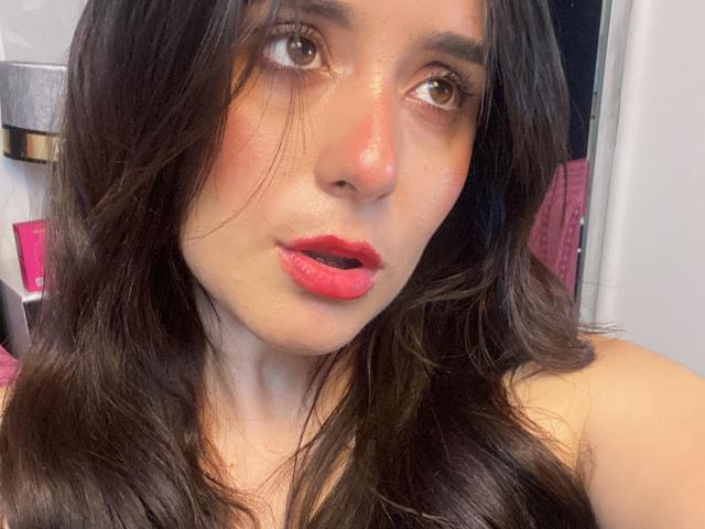 VickyMegan with dark brown wavy hair, red lipstick, and slightly parted red lips, gazing to the side with brown eyes, showing a bit of her teeth. Her skin is shiny.