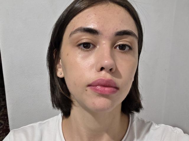 RebbeccaLipinsky has a sweaty face, brown eyes, pink lips, small nose, brown bob haircut, white shirt, and a few moles. Her face is slightly shiny and she looks serious.