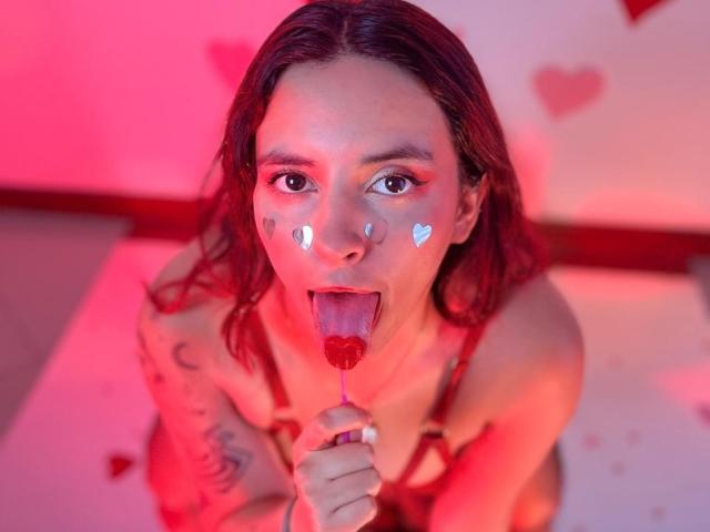 LaiaJonnes, avec des cheveux roux, des autocollants en forme de cœur sur les joues, la langue tirée, léchant un bonbon en forme de cœur rouge, des tatouages sur les bras, un éclairage rose et rouge, nue, tenant le bonbon devant elle..
