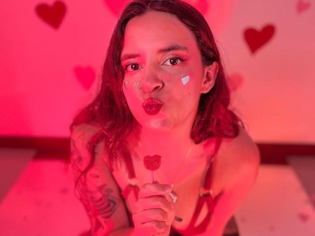 LaiaJonnes, avec du rouge à lèvres, un maquillage en forme de cœur et un bras tatoué, tenant un bonbon en forme de cœur scintillant, faisant la moue avec un éclairage rose-rouge, portant de la lingerie rouge à bretelles..