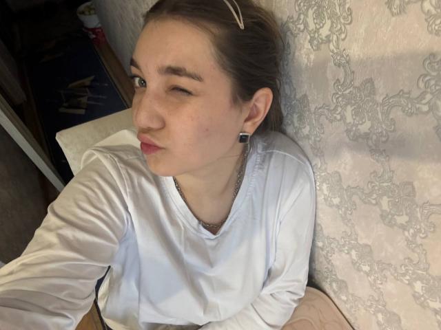 MichikoVivona with winking eye, wearing a white long-sleeve shirt, has a chain necklace, and small black ear gauges, sitting against a patterned wall.