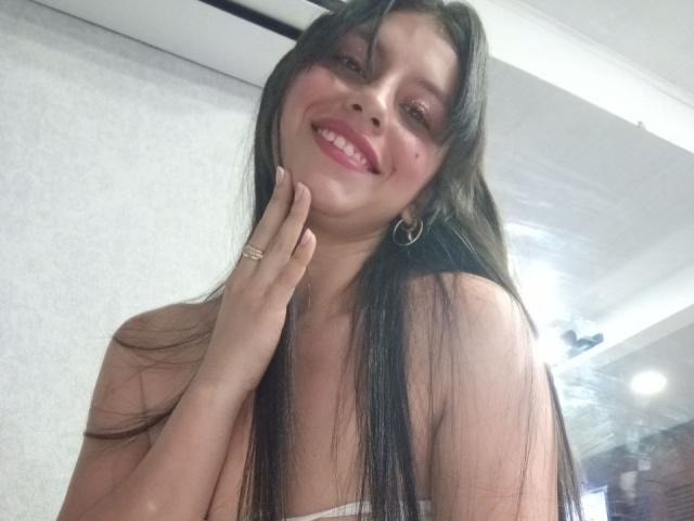 MadissonFontaine with long black hair, smiling with pink lipstick, holding her chin, wearing a strapless top, showing her tits, wearing a gold ring and hoop earrings.