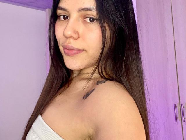 SophiaMarx with long brown hair, light brown eyes, and a small tattoo on her shoulder. She's wearing a white strapless top, showing her bare shoulders and small tits.