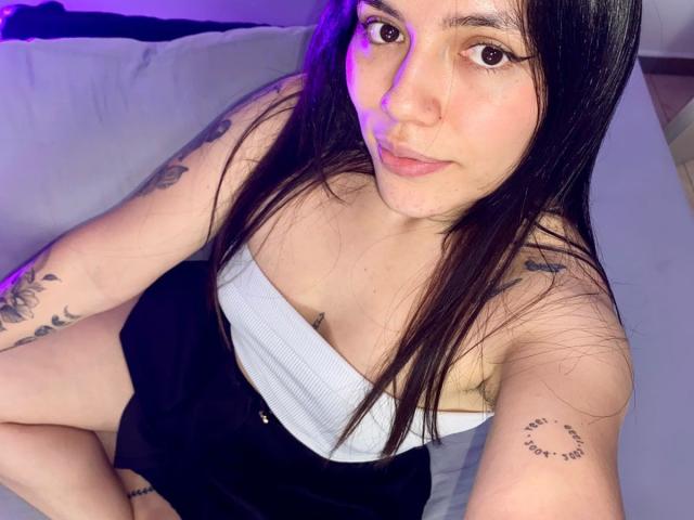 SophiaMarx with long brown hair, white tube top, black shorts, tattoos on arms, sitting on bed, purple light, looking at camera, slight smile, visible nose ring, soft lighting.
