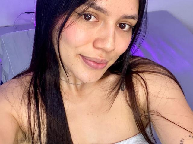 SophiaMarx with long black hair, brown eyes, light skin, small nose, subtle smile, and visible freckles, wearing a white strapless top, showing her tits.