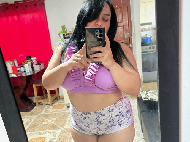 Viviana Thomson, avec ses cheveux noirs, son débardeur de sport violet et son short blanc à motifs floraux, tenant un téléphone, prenant un selfie devant un miroir, montrant ses seins et ses fesses..