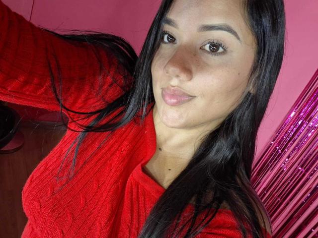 KinkyDaycaree, avec de longs cheveux noirs, des yeux marron et un sourire mignon, portant un haut tricot rouge, laissant entrevoir ses petites seins, et un petit anneau au nez, sur un mur rose..