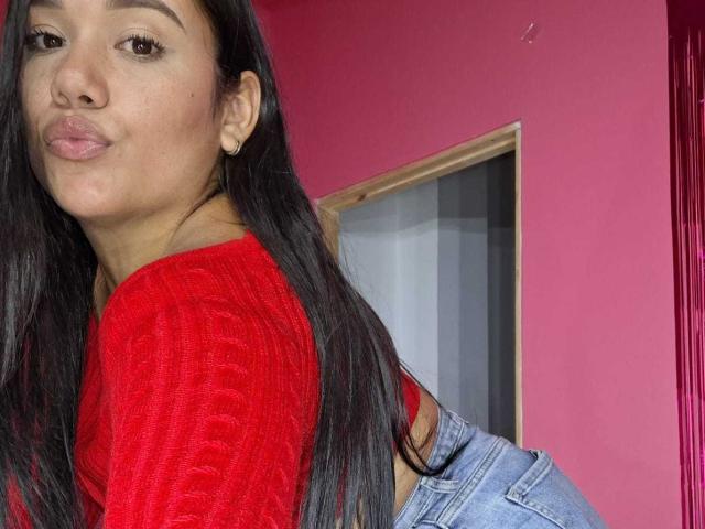 KinkyDaycaree, avec de longs cheveux noirs, un pull rouge en tricot et un jean bleu, faisant la moue, portant une petite boucle d'oreille dorée, debout devant un mur rose..