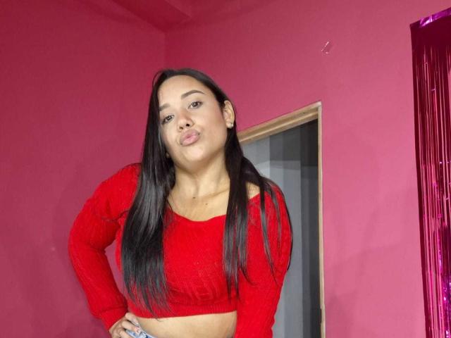 KinkyDaycaree, avec de longs cheveux noirs, portant un crop top rouge à manches longues, les lèvres pincées, debout devant un mur rose, avec une expression confiante. Les seins sont légèrement visibles sous le haut..