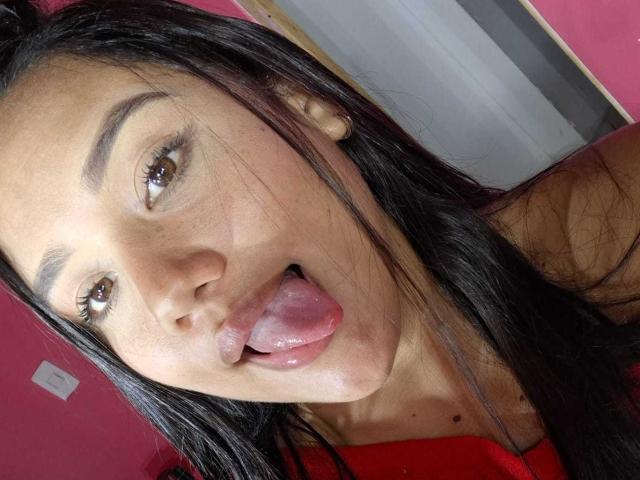 KinkyDaycaree a de longs cheveux noirs, des yeux marrons, et elle sort sa langue rose et mouillée, la bouche légèrement ouverte. Elle porte une chemise rouge..