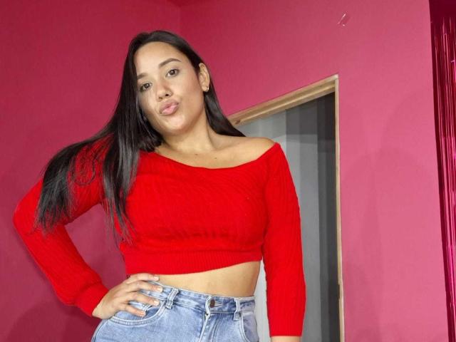 KinkyDaycaree, avec de longs cheveux noirs, un haut rouge à une épaule et un jean bleu, debout dans une pièce aux murs roses, faisant la moue et posant une main sur sa hanche..