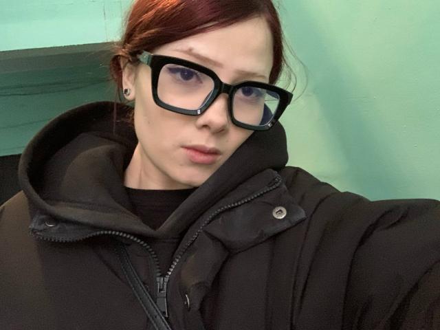 PatricaCitino with red hair, black glasses, and a black jacket, showing a pouty face, has small tits and a nose ring, looking slightly annoyed.