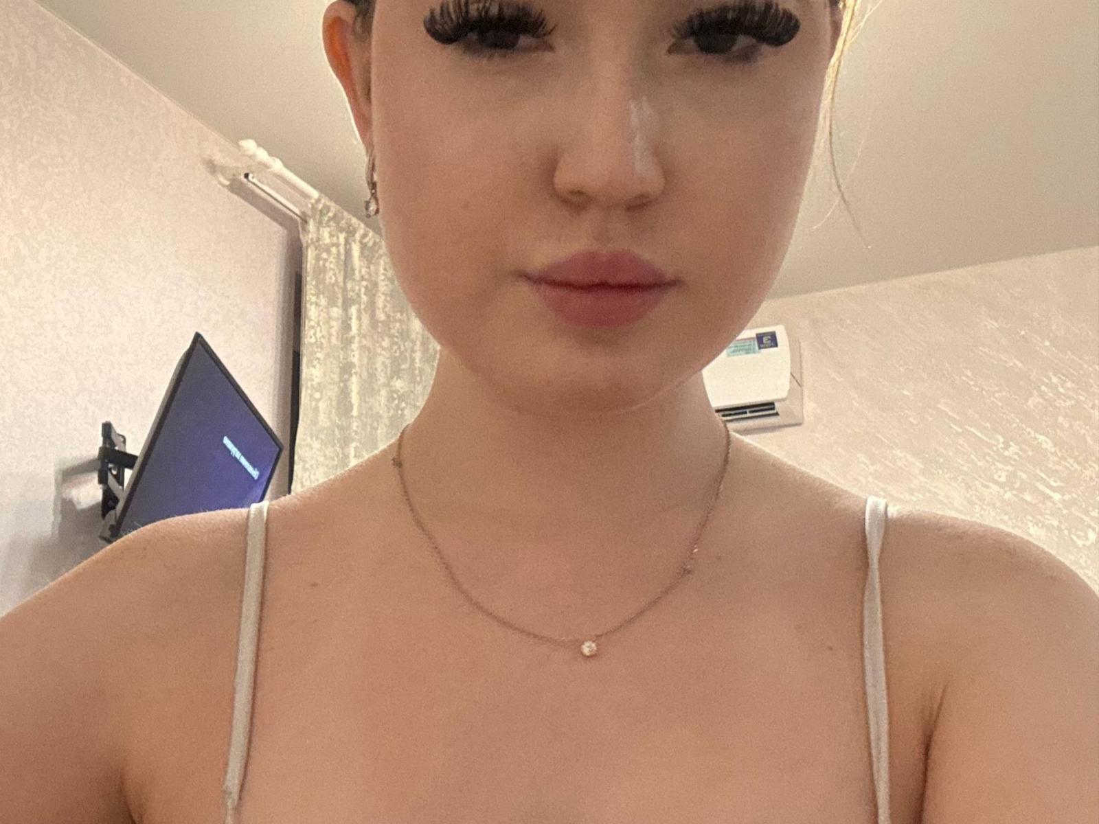 NadiaDeanhardt with light skin, full lips, and long dark eyelashes wearing a white spaghetti strap top, necklace with small pendant, standing in a room with a TV.