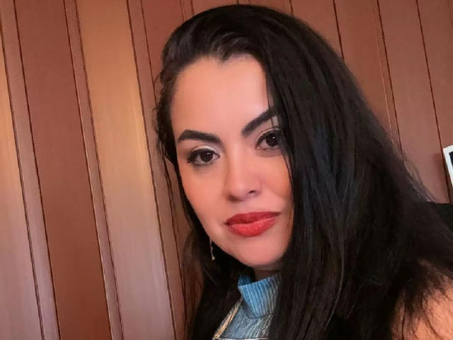 Charlotte Devereaux hat dunkles, glänzendes Haar, roten Lippenstift und trägt einen blauen Rollkragenpullover. Sie hat dicke Augenbrauen und dezentes Make-up und blickt mit einem leichten Lächeln in die Kamera..